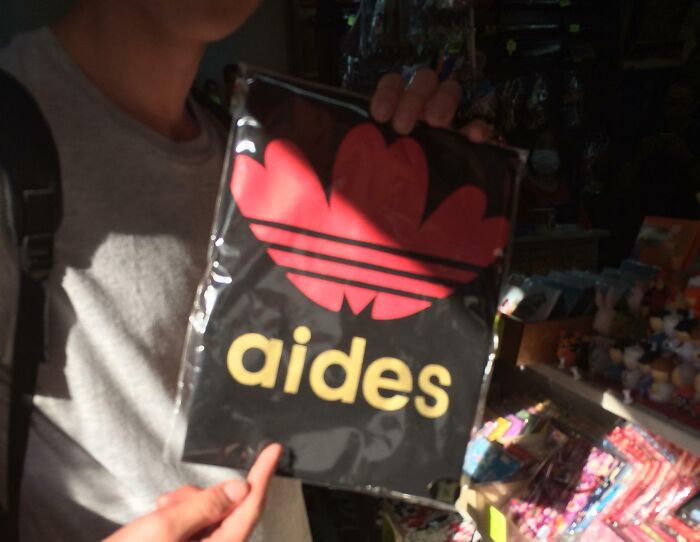 Person holding a hilariously bad T-shirt design resembling a famous logo with the word aides in a crowded shop.