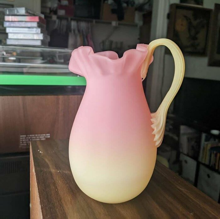 Frosted pink and yellow glass pitcher with ruffled edge and ornate handle displayed on wooden surface.