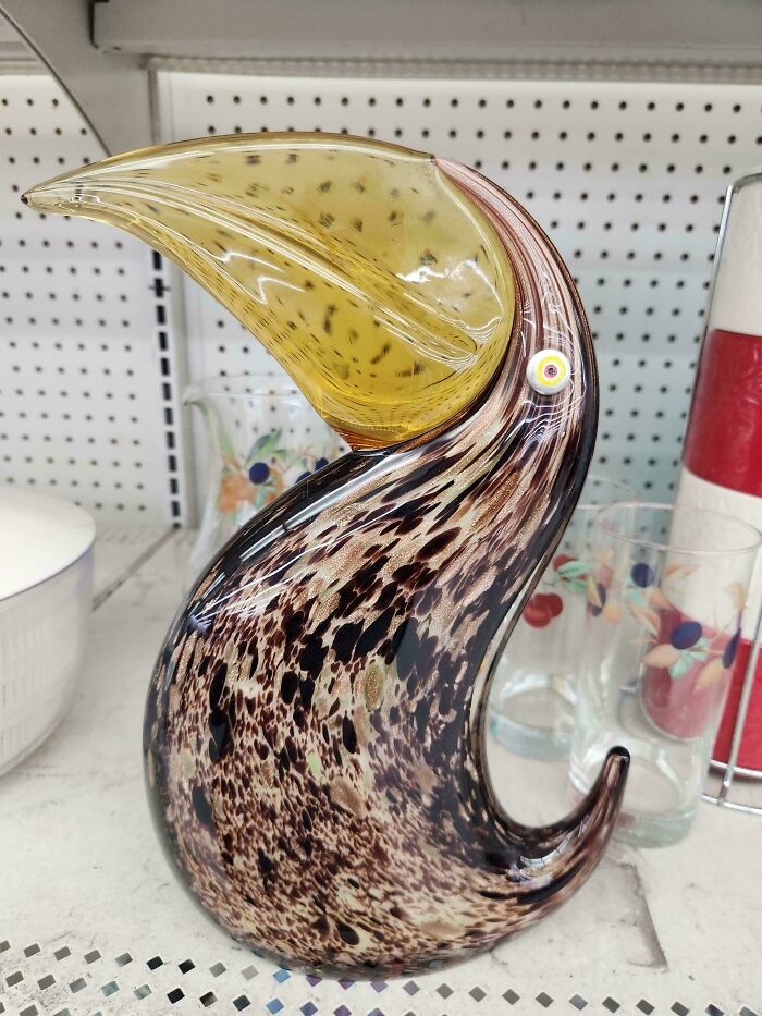 Handcrafted glass bird sculpture with a large yellow beak and speckled brown body displayed on a shelf among glassware.