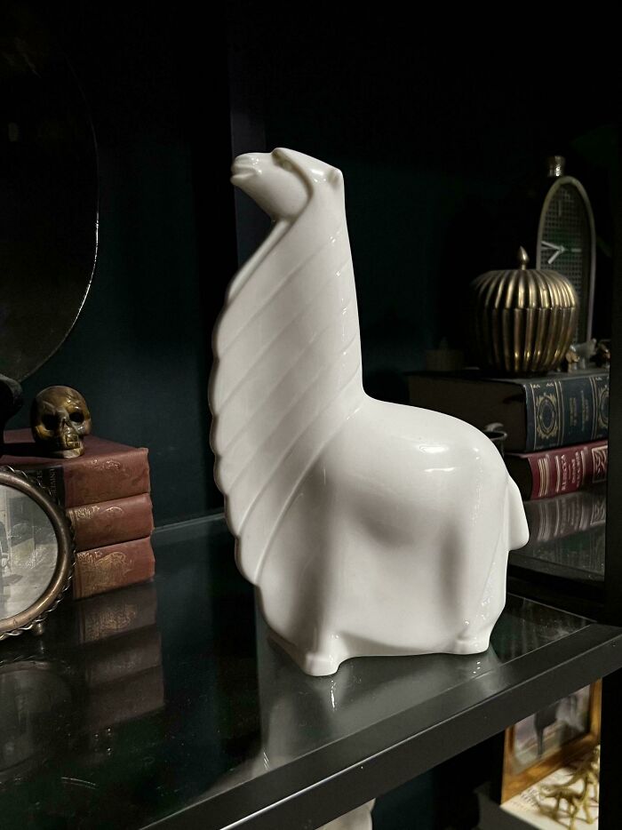 White glass llama figurine with textured wings displayed on a reflective shelf among vintage decor items.