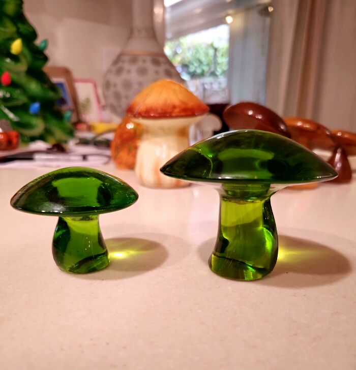 Two small green glass mushroom figurines displayed on a table among creative glass items for home decor.