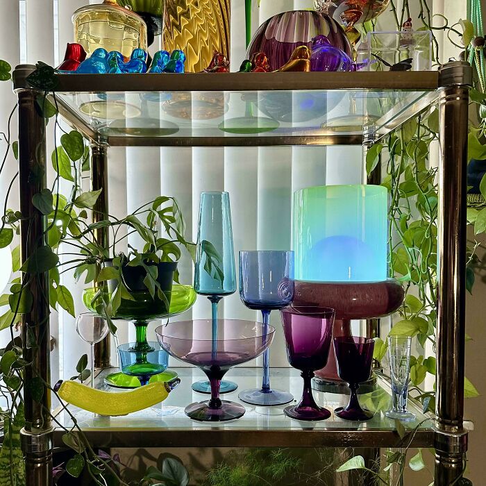 Colorful and creative glass items displayed on a metal and glass shelf surrounded by green houseplants.