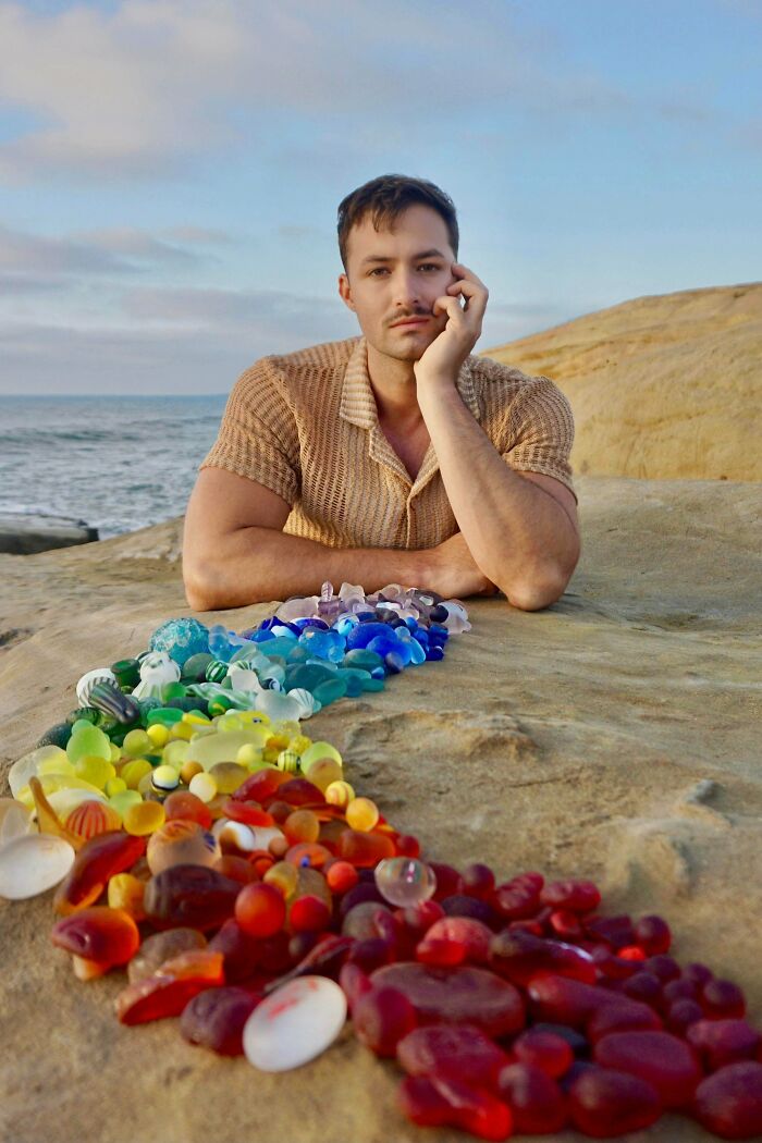 Man leaning on rock by ocean displaying colorful creative glass items arranged in a rainbow pattern outdoors at sunset