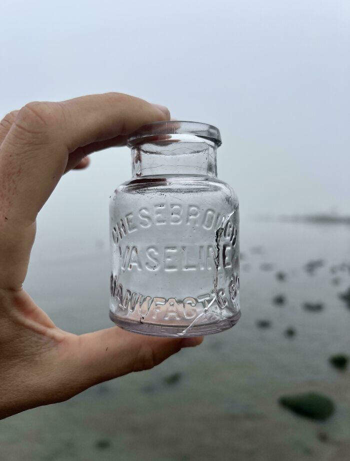 Hand holding a cracked vintage glass vaseline jar found near a foggy shoreline, a cool and creative glass item for home.