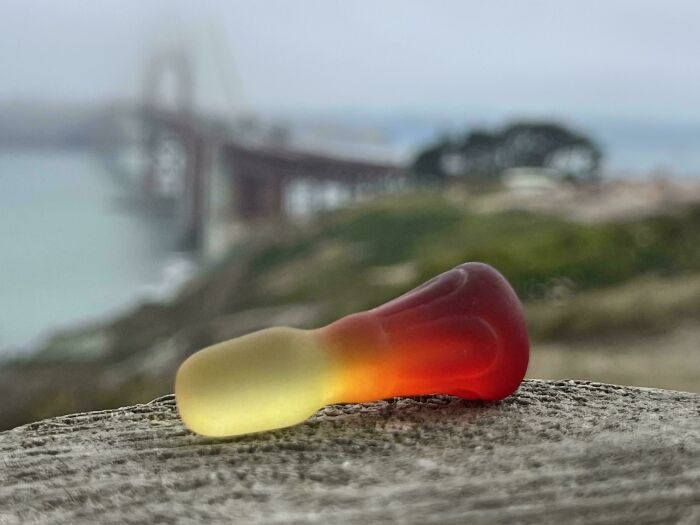 Colorful glass art piece shaped like a gummy candy displayed outdoors near a blurred bridge and greenery background.