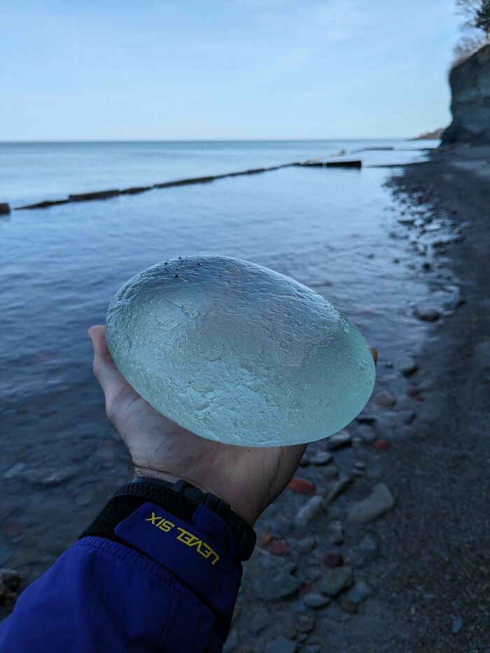 Hand holding a smooth, clear glass item found by the rocky shoreline with ocean water and cliffs in the background.