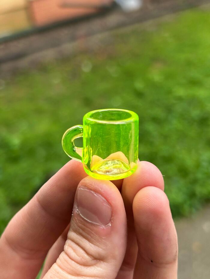 Hand holding a small, bright green creative glass item shaped like a miniature mug outdoors with blurred grass background