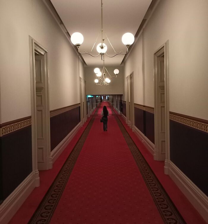 Person standing in a long hotel hallway with red carpet and chandeliers creating an unedited photo that messes with your brain.