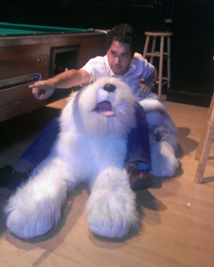 Man sitting on the floor behind a large, fluffy dog plush toy, pulling off a creative and hilarious pose perfectly.