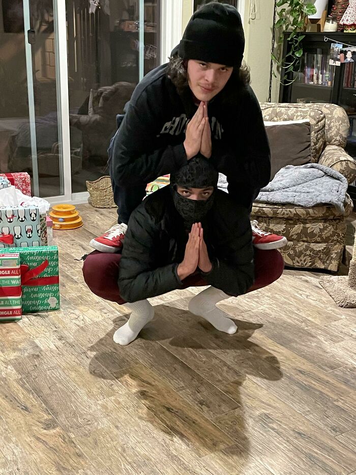 Two people striking creative and hilarious poses indoors, balancing on each other with hands in prayer position.