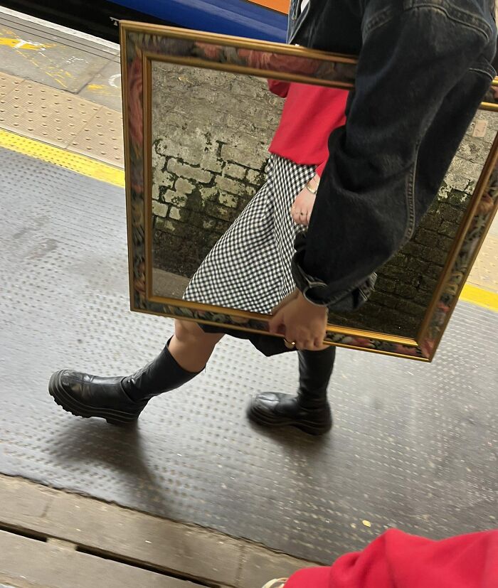 Person holding a framed mirror reflecting a brick wall and checkered skirt, creating an optical illusion to mess with your brain.