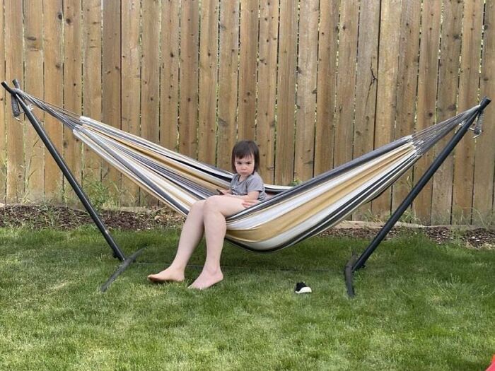 Child sitting awkwardly on a hammock with legs stretched out, one foot on the ground, in an outdoor backyard setting.