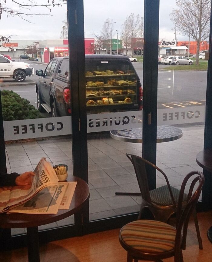 Pickup truck parked outside coffee shop with dessert display inside, a visually confusing unedited photo likely to mess with the brain.