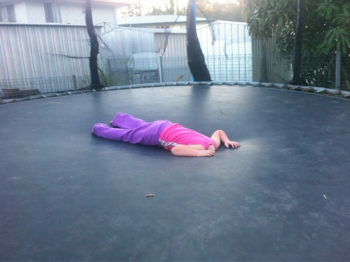 Child lying face down on a trampoline, wearing purple pants and a pink shirt, creating a brain-messing visual effect.