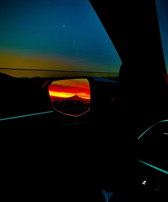 Sunset reflection in a car side mirror with vibrant orange and red hues against a dark blue sky, an unedited photo messing with perception.