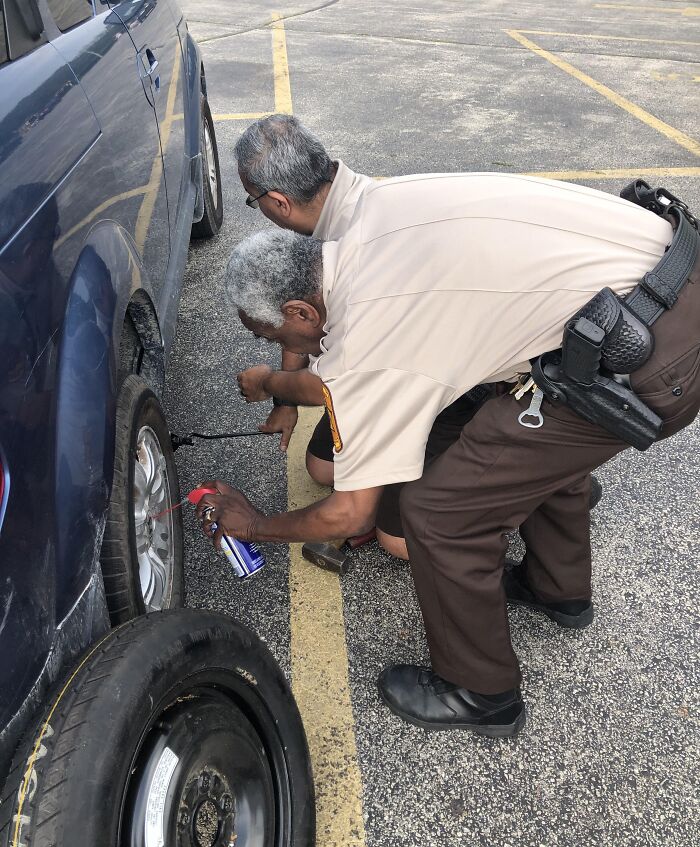 Two men fixing a flat tire on a parked car in a parking lot in a photo that might mess with your brain.