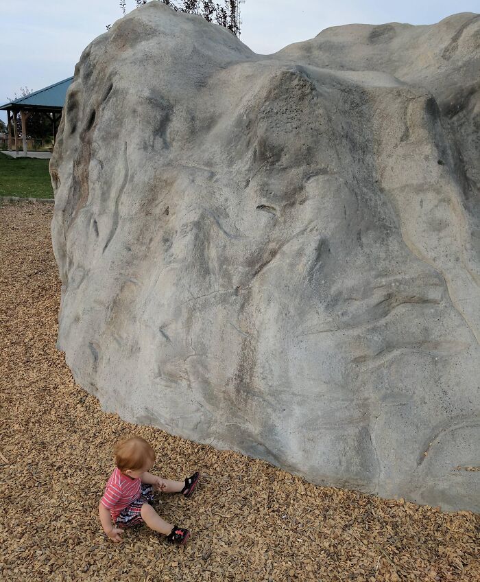 Toddler sitting on wood chips next to a large rock with unedited photo textures that might mess with your brain.