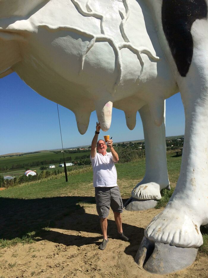 Man standing under a giant cow statue, pretending to milk it, capturing the essence of the US state in this picture.