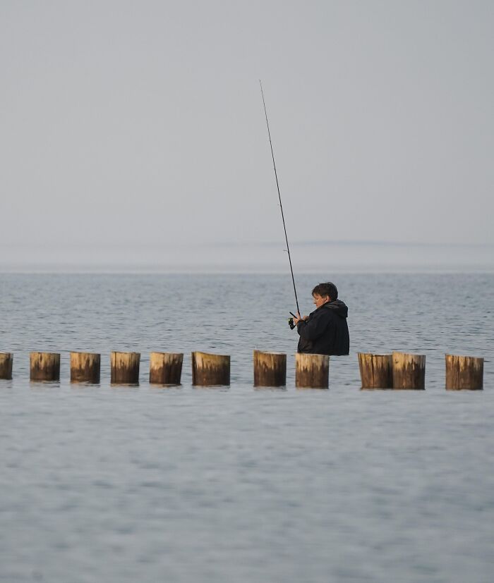 Person fishing while standing on submerged wooden posts in calm water creating a mind-bending unedited photo effect.