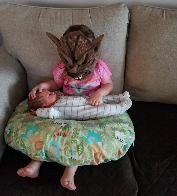Toddler wearing a wolf mask holding a baby on a colorful pillow, capturing a funny moment of sibling rivalry.