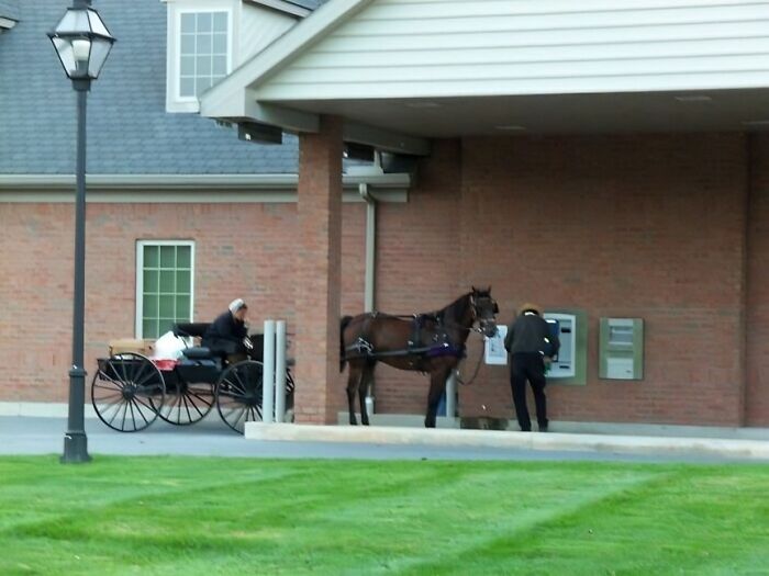Horse-drawn carriage beside brick building with person at ATM, capturing the essence of rural US states in a classic scene.