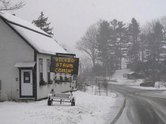 Snow-covered rural road and house with a digital sign reading wicked stawm in a scene capturing the essence of US states.