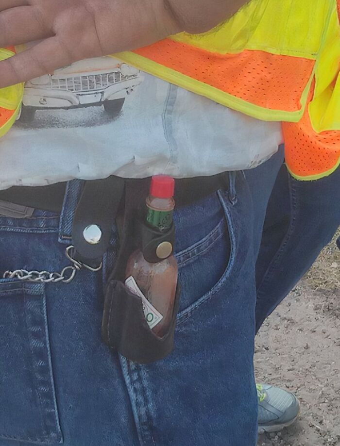 Person wearing a safety vest and jeans with a hot sauce bottle and cash tucked into their belt pouch in a rural setting.
