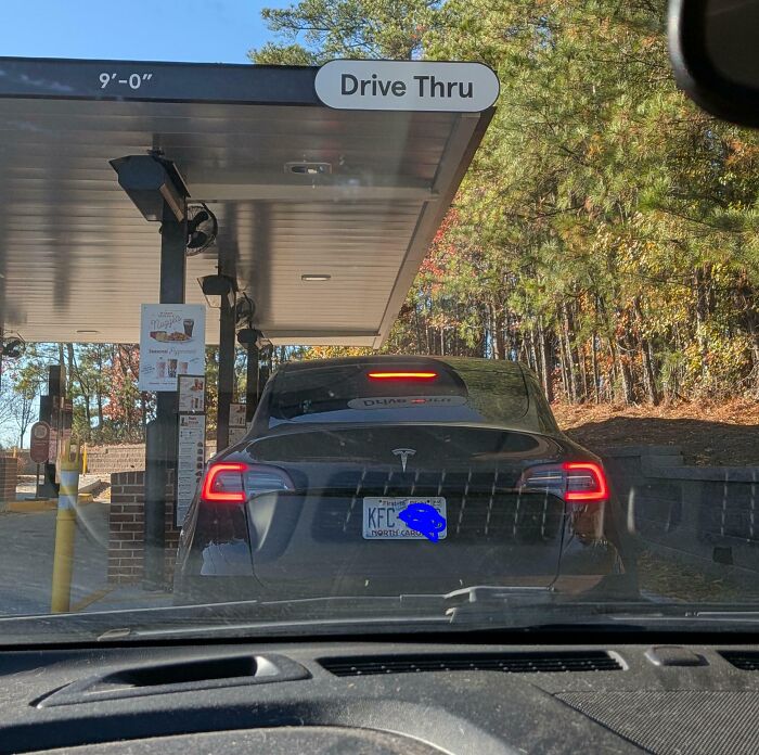 Tesla electric car with a customized KFC license plate waiting in a drive-thru surrounded by autumn trees.
