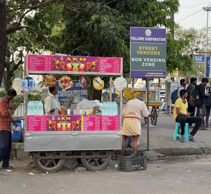Street vendors gathered near a juice bar cart under a Vellore Corporation non vending zone sign outdoors.