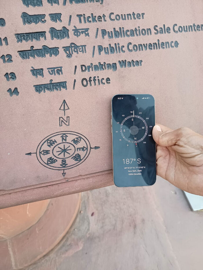 A person holding a smartphone with a compass app showing South, pointing to a North arrow on a sign, a funny you had one job fail.