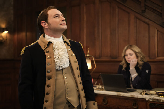 Actor dressed in period costume portraying William, with a woman working on a laptop in a wood-paneled room, related to Invincible voice actors.