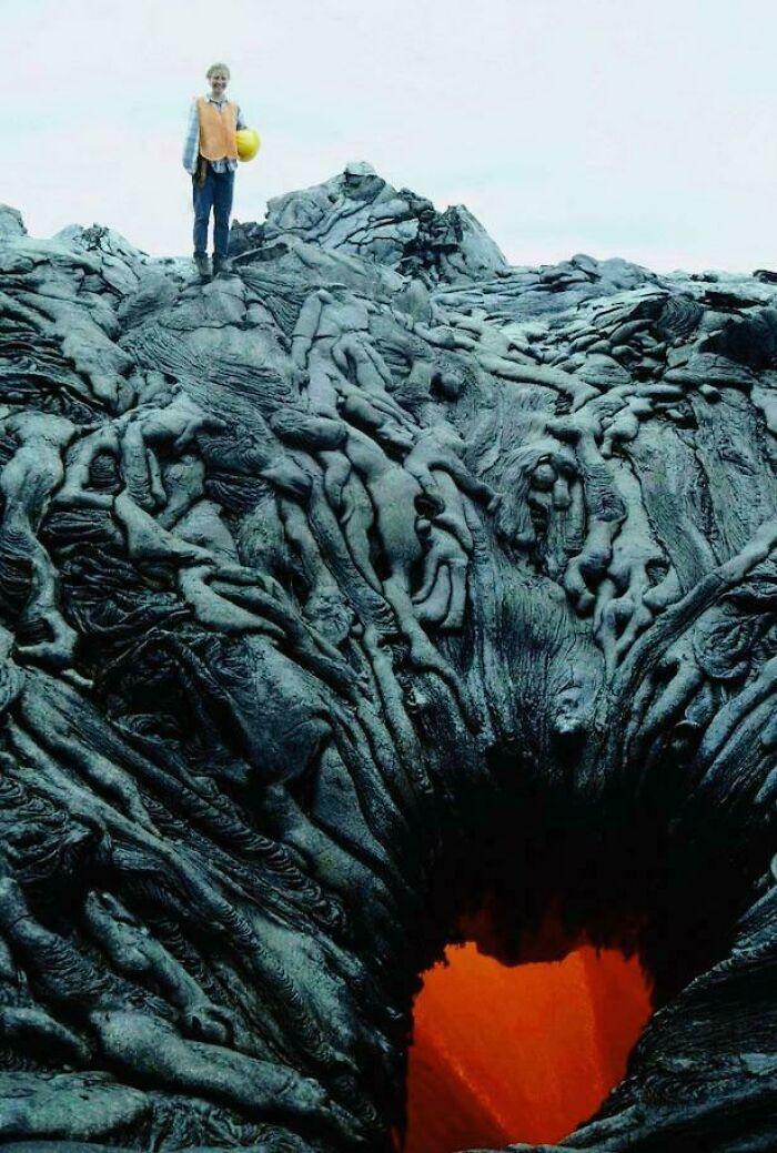 A person stands on a creepy, hardened lava flow that resembles entangled bodies, looking down into a glowing lava pit.