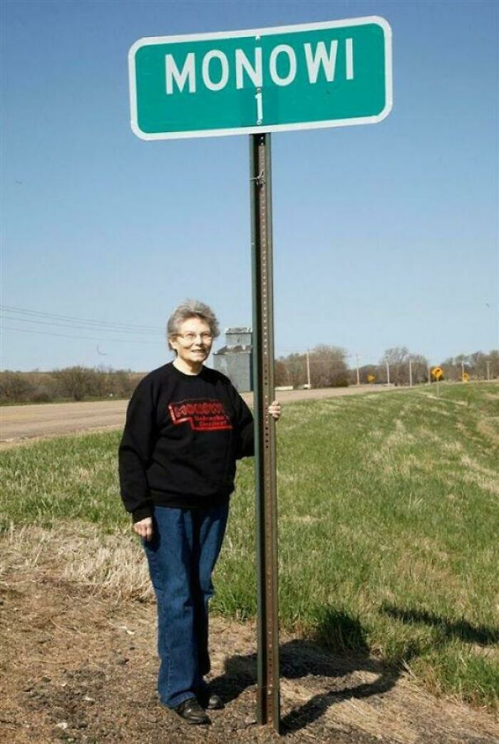 Elderly woman standing by a Monowi town sign, showcasing an interesting vault of bizarre and cool world oddities.