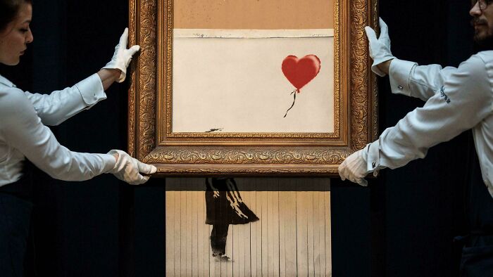 Two gallery workers carefully lift a famous street art painting featuring a red heart balloon, representing interesting vault stories.