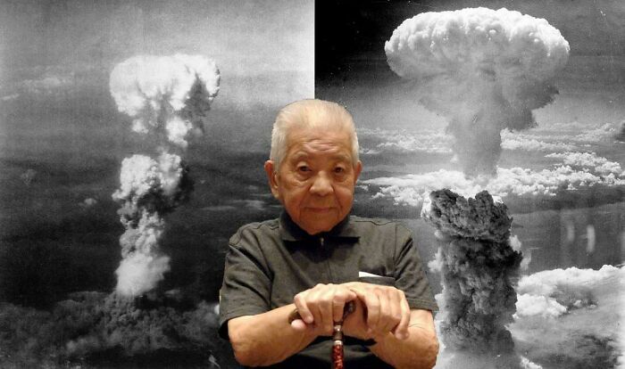 Elderly man sitting with hands on a cane in front of historic mushroom cloud photos, capturing interesting vault stories.