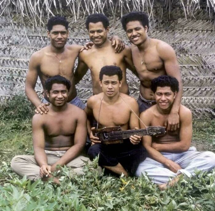 Group of six shirtless men sitting and standing outdoors with a handmade guitar, showcasing interesting vault stories and oddities.