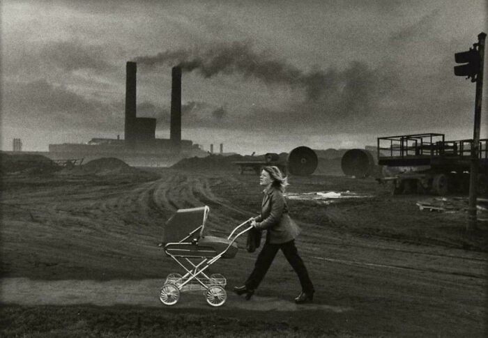 Woman pushing stroller in smoky industrial urban hell area with factory chimneys
