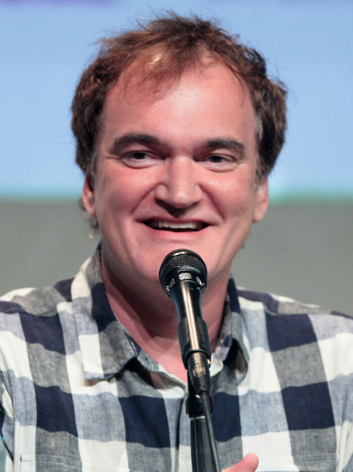 Quentin Tarantino speaking into microphone at an event, showcasing his bio and career highlights in filmmaking.