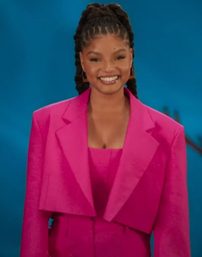 Halle Bailey smiling in a vibrant pink outfit against a blue background, highlighting her bio and career highlights.
