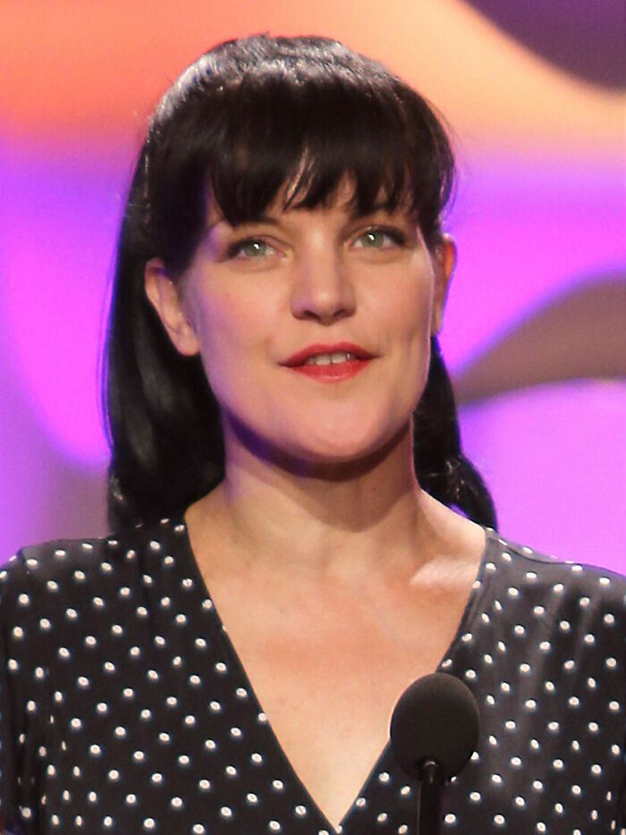 Pauley Perrette speaking on stage with a microphone, wearing a black polka dot top in a bright setting.