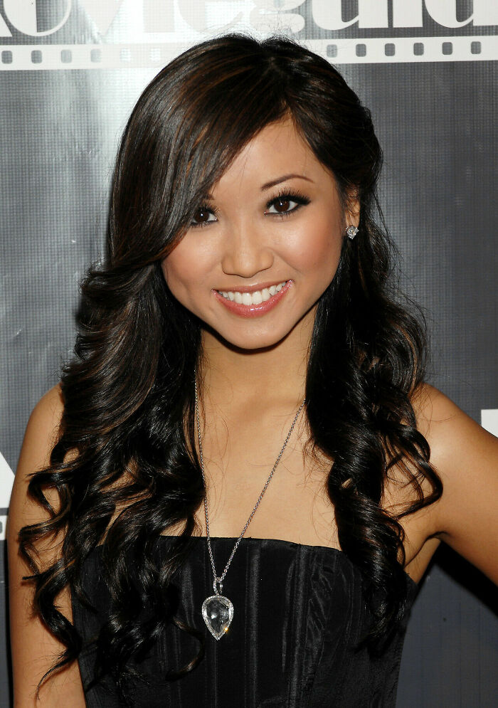 Brenda Song smiling at event with long curly hair, wearing a black dress and a heart-shaped pendant necklace.