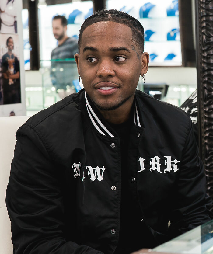 London on da Track wearing a black New York jacket, sitting indoors with braided hair and earrings, looking to the side.