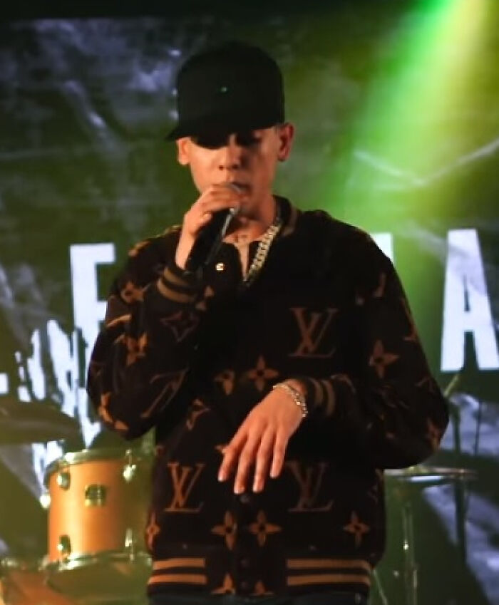Natanael Cano performing on stage wearing a black cap and Louis Vuitton jacket under green stage lights.