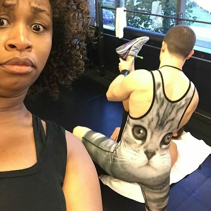 Woman making a shocked face at the gym while a man wears a workout outfit with a large cat face print.