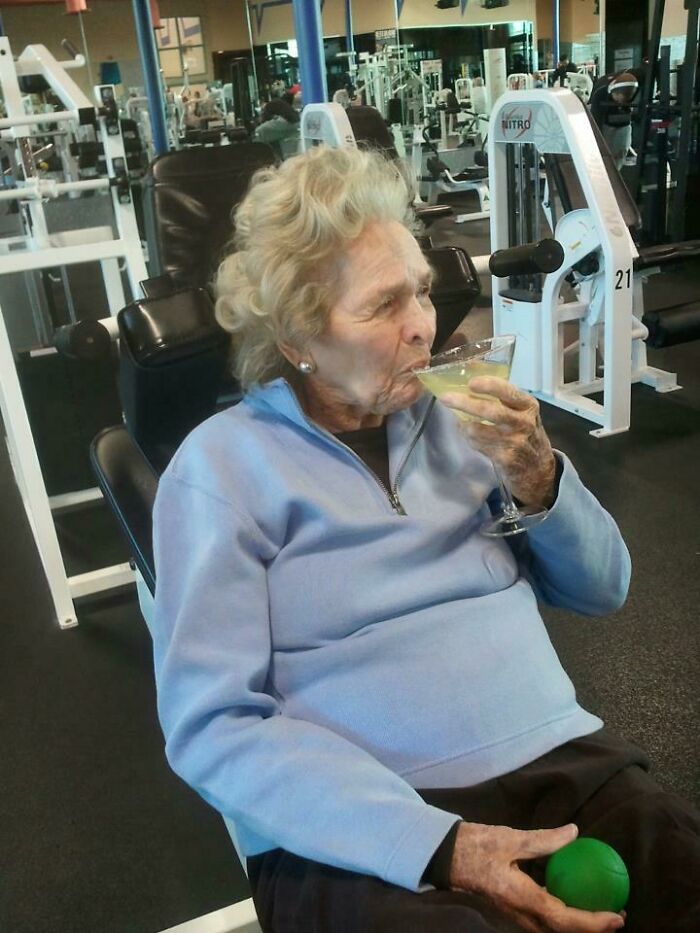 Elderly woman sitting in gym workout chair sipping cocktail, holding green exercise ball in unusual gym moment.