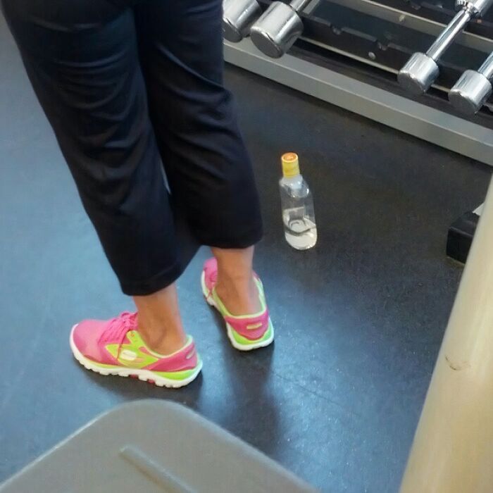 Person wearing pink and green sneakers standing near weights in a gym, a clear bottle placed on the floor nearby.