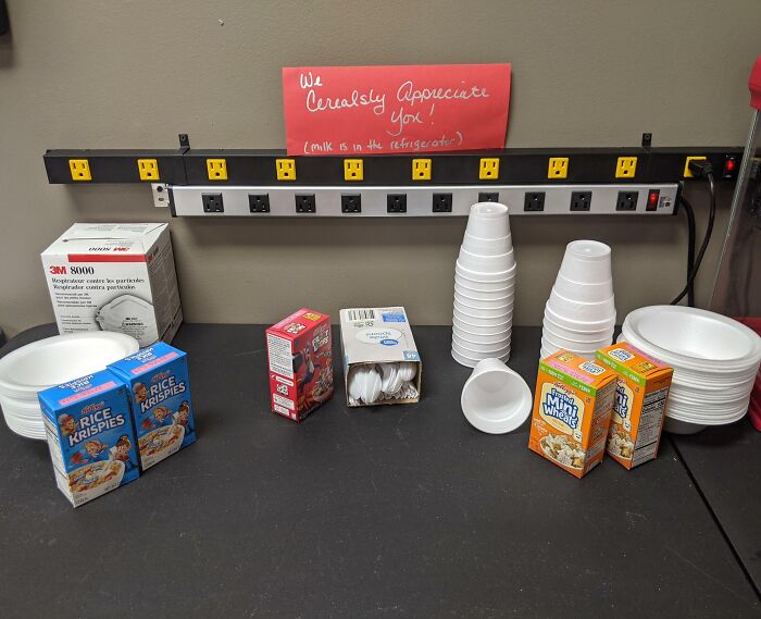 Workplace break room with cereal boxes, disposable plates, cups, and a humorous appreciation note on the wall.