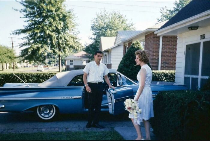 A candid old photo shows a couple beside a blue vintage car, capturing a moment from generations ago.