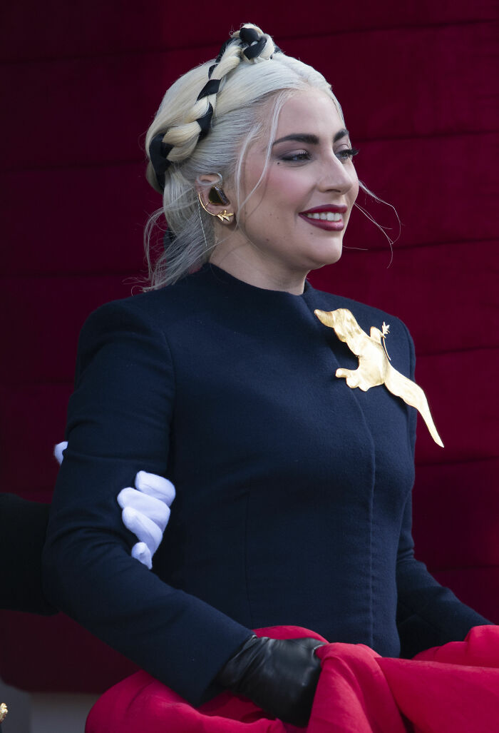 Lady Gaga smiling, wearing a braided hairstyle and black outfit with gold detail, highlighted in her bio and career.