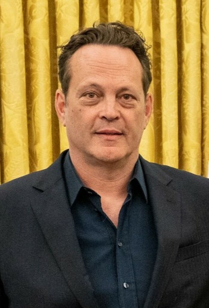 Vince Vaughn wearing a dark suit standing in front of gold curtains at a formal event, showcasing his career highlights.